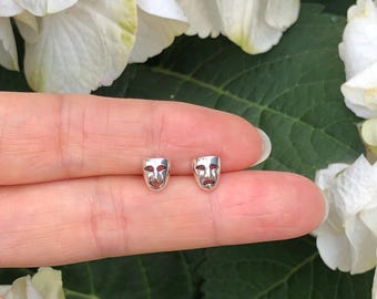 May include: A pair of silver earrings featuring a small, detailed design of a theater mask. The earrings are shown on a person's finger against a backdrop of white flowers.
