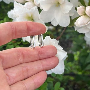 May include: A pair of small, silver hoop earrings held in a hand. The earrings have a rectangular shape with a subtle design. The background features white flowers and green foliage.