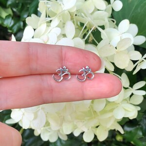 May include: A pair of small, silver-colored Om symbol earrings. The earrings are held up against a backdrop of white and green flowers. The Om symbol is a sacred sound and spiritual icon in Hinduism.