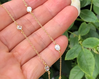 May include: A gold chain necklace with small round clear stones. The necklace is on a hand with a green leafy background.