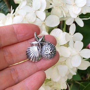 May include: Silver charm featuring a seashell, a starfish, and a sand dollar. The charm is held in a hand against a background of white flowers.