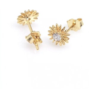 Peut inclure: Une paire de boucles d'oreilles dor&eacute;es. Chaque boucle d'oreille a la forme d'un &eacute;clat de soleil stylis&eacute; avec une petite pierre pr&eacute;cieuse transparente au centre. Les boucles d'oreilles sont pr&eacute;sent&eacute;es sur fond blanc.