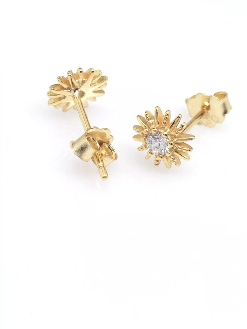 Peut inclure: Une paire de boucles d'oreilles dor&eacute;es. Chaque boucle d'oreille a la forme d'un &eacute;clat de soleil stylis&eacute; avec une petite pierre pr&eacute;cieuse transparente au centre. Les boucles d'oreilles sont pr&eacute;sent&eacute;es sur fond blanc.