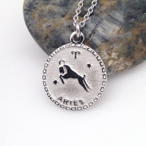 May include: Silver necklace with a round pendant featuring the zodiac symbol for Aries. The pendant has a textured surface and the word "Aries" is inscribed around the edge.