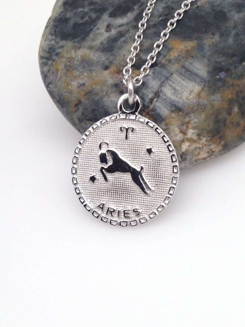 May include: Silver necklace with a round pendant featuring the zodiac symbol for Aries. The pendant has a textured surface and the word "Aries" is inscribed around the edge.