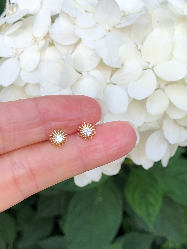 Peut inclure: Une paire de boucles d'oreilles dor&eacute;es. Chaque boucle d'oreille pr&eacute;sente un motif de soleil avec une pierre pr&eacute;cieuse claire au centre. Les boucles d'oreilles sont petites et d&eacute;licates. Fond flou avec des fleurs blanches.