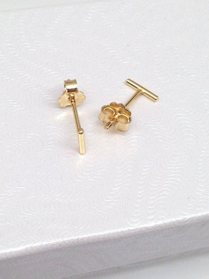 K&ouml;nnte beinhalten: Ein Paar goldfarbene Ohrstecker mit einem geraden Stabdesign. Die Ohrringe haben einen Stift und einen Schmetterlingsverschluss.
