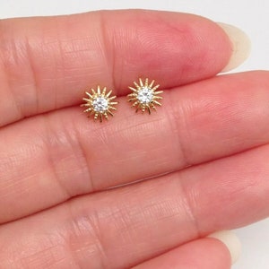 Peut inclure: Une paire de boucles d'oreilles &agrave; clous dor&eacute;es. Chaque boucle d'oreille pr&eacute;sente un motif de soleil avec une pierre ronde et transparente au centre. Les boucles d'oreilles sont petites et d&eacute;licates.