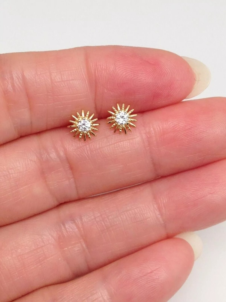 Peut inclure: Une paire de boucles d'oreilles &agrave; clous dor&eacute;es. Chaque boucle d'oreille pr&eacute;sente un motif de soleil avec une pierre ronde et transparente au centre. Les boucles d'oreilles sont petites et d&eacute;licates.