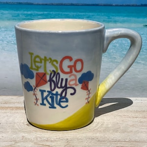 May include: A white ceramic mug with a yellow, blue, and pink design. The mug has a handle and says "Let's Go Fly a Kite" on the front.