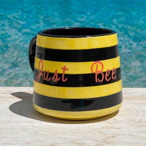May include: A ceramic mug with a black handle and a black interior. The mug features alternating horizontal yellow and black stripes. The words "Just Bee" are written in pink script across the front of the mug.