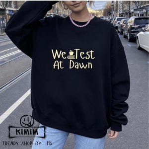 Może przedstawiać: Czarna bluza z napisem "We Test At Dawn" w żółtym kolorze. Projekt zawiera jabłko i książki. Bluza pochodzi z kolekcji "Trendy Shop by MS".