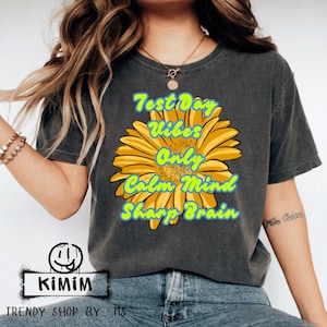 Peut inclure: T-shirt gris foncé avec un grand motif de fleur jaune. Le motif floral contient le texte "Test Day Vibes Only Calm Mind Sharp Brain" en bleu et vert. Le t-shirt a aussi le texte "KIMIM" et "TRENDY SHOP BY MS".