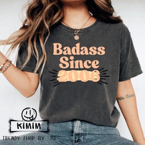 Puede incluir: Camiseta gris oscuro con el texto "Badass Since 2006" en melocotón y amarillo. La camiseta tiene un aspecto vintage y desgastado. La parte inferior de la camiseta tiene un logotipo que dice "KIMIM" y "TRENDY SHOP BY MS".