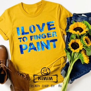 May include: Mustard yellow t-shirt with the text "I LOVE TO FINGER PAINT" in blue brushstrokes. The shirt is paired with blue jeans, sunflowers, and brown leather boots. A logo with a smiley face and the text "KIMIM" is at the bottom.