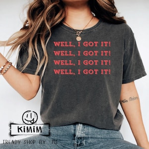 May include: Dark gray t-shirt with the text "WELL, I GOT IT!" in red. The shirt has a vintage, worn look. The image also includes the shop's logo and the text "TRENDY SHOP BY MS".