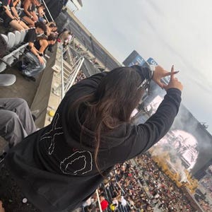 May include: A black hooded sweatshirt with white embroidered "XO" design on the back. The person is at a concert, with a large crowd and stage visible in the background. The person is making a frame with their hands.