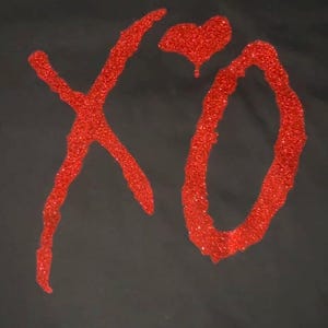 Puede incluir: Camiseta negra con un diseño de purpurina roja. El diseño presenta las letras "XO" con un corazón encima de la "O". Las letras y el corazón son de purpurina roja.
