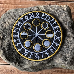 May include: A circular black patch with a gold border and white and gold embroidered design. The design features a central starburst pattern with seven circles, each containing a moon phase. The outer ring has runes.