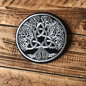 May include: A silver embroidered patch featuring a tree of life design on a black background. The tree is encircled by a Celtic knot.