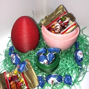 Könnte beinhalten: Ein rotes, strukturiertes Ei und ein rosa eiförmiger Behälter, gefüllt mit verpackten Süßigkeiten, liegen auf grünem Ostergras. Mehrere blau-weiß verpackte Süßigkeiten und goldene Süßigkeitenverpackungen sind um die Behälter herum verstreut. Ein kleiner grüner Behälter enthält weitere Süßigkeiten.