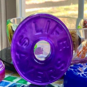 May include: A vibrant purple, circular object with the words "LIFE SAVERS" embossed around the edge. The object has a hole in the center. Other colorful objects are in the background, including a pink and blue item.