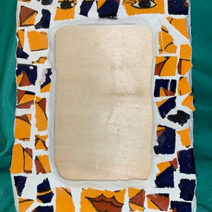 May include: A mosaic picture frame with a wood backing. The frame is made of small, broken pieces of tile in shades of orange, blue, and white. The frame features a stylized face with eyes, eyebrows, and a mouth.