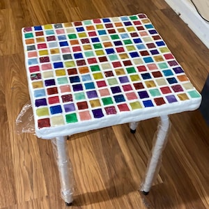 May include: A small, white table with a colorful mosaic tile top. The tiles are arranged in a grid pattern and are made of glass. The table has three legs and is wrapped in plastic.