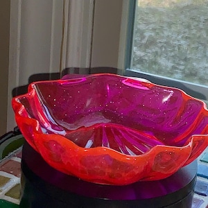 May include: A decorative, translucent bowl with a scalloped edge. The bowl is a vibrant mix of red and magenta, with a glittery interior. The bowl is sitting on a dark surface.