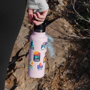 May include: A light pink water bottle with a black lid, covered in colorful stickers featuring mountain and nature scenes. The stickers include text such as "Zion", "Acadia", and "Lake Clark".