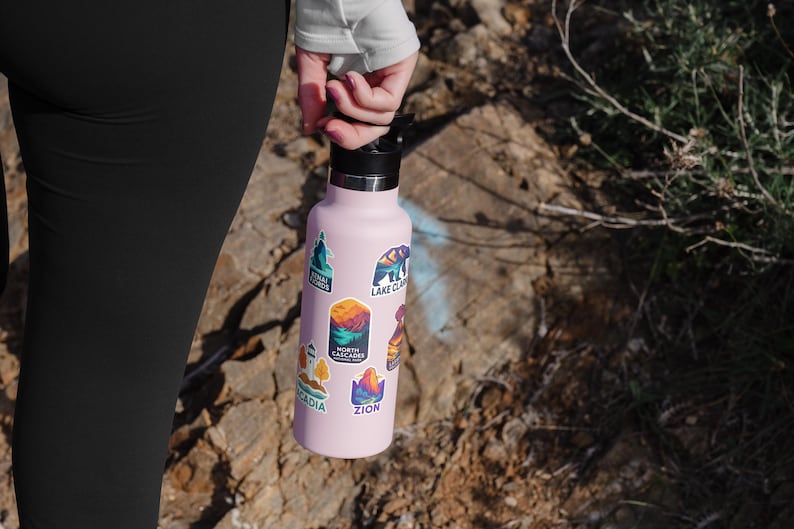 May include: A light pink water bottle with a black lid, covered in colorful stickers featuring mountain and nature scenes. The stickers include text such as "Zion", "Acadia", and "Lake Clark".
