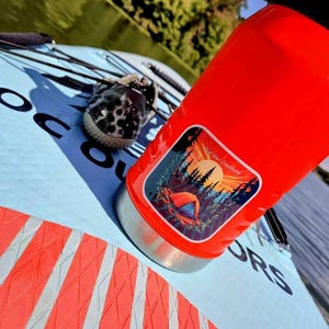 May include: A bright red insulated water bottle with a camping-themed sticker, resting on a light blue paddleboard with orange and white accents. A pair of black and white patterned water shoes are also on the paddleboard.