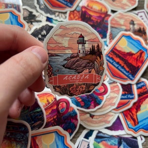 May include: A collection of stickers featuring scenic illustrations. One sticker depicts a lighthouse on a rocky coast with the text "ACADIA National Park ME". Other stickers show mountain landscapes and national park scenes.