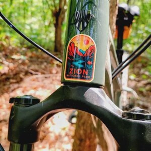 May include: A dark green bicycle frame featuring a black head tube badge and a colorful sticker depicting a landscape with red rock formations, a blue river, and the text Zion National Park in bold orange letters.