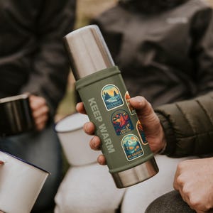 May include: A green and silver insulated thermos with the text "KEEP WARM" printed vertically. The thermos has several colorful stickers featuring national park designs. The thermos is held by a person.