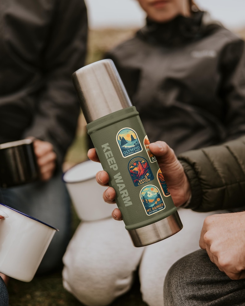 May include: A green and silver insulated thermos with the text "KEEP WARM" printed vertically. The thermos has several colorful stickers featuring national park designs. The thermos is held by a person.