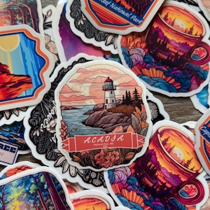 May include: A collection of colorful stickers featuring nature scenes. Designs include a lighthouse with "Acadia National Park, ME" text, a mountain landscape, and a sunset over a forest. The stickers have a white border.
