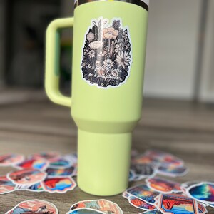 May include: A light green insulated tumbler with a handle, a white lid, and a straw. A sticker with a floral design and the text "In a field of roses, be a wildflower" is on the tumbler. Assorted stickers with landscape designs are scattered around the tumbler.