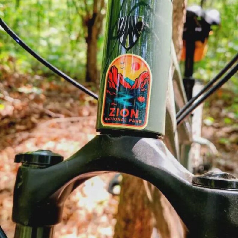 May include: Close-up of a green bicycle frame with a Zion National Park sticker. The sticker features a landscape with red rock formations, a river, and a sunset. The bike's fork and handlebars are black.