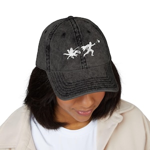 May include: A charcoal black baseball cap with a distressed, washed-out finish. The front features a white graphic print of a figure wearing a cape throwing a ball, while a creature with wings and a crown holds a fishing rod with a small ring.