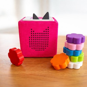 May include: A bright pink, cube-shaped audio player with black cat ears and a speaker grille in the shape of a cat face. A stack of colorful flower-shaped blocks sits next to the player, with a red and orange block nearby.