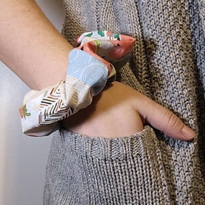 Puede incluir: Un scrunchie de tela colorido con un diseño de patchwork que presenta remolinos, patrones geométricos y motivos florales. El scrunchie está atado alrededor de la muñeca de una persona.