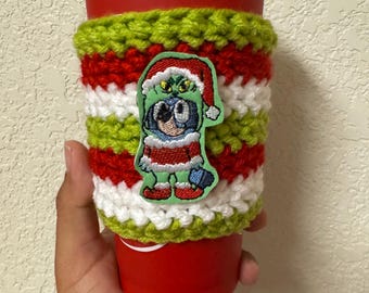 May include: A red disposable coffee cup with a white lid, wrapped in a handmade crochet cozy. The cozy features green, red, and white stripes, and a patch of a cartoon character in a Santa hat. The cozy is designed to insulate the cup.