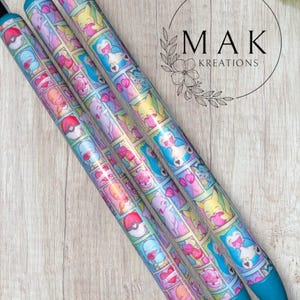 May include: Four pens with a colorful cartoon design featuring various characters. The pens have a blue tip and a black top. The design includes a variety of colors, including pink, yellow, and blue. The pens are arranged diagonally on a light wood surface.