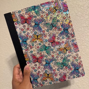 May include: A notebook with a black spine and a cover featuring a repeating pattern of colorful butterflies and small flowers on a light beige background. The butterfly wings have a holographic finish that reflects light with various rainbow colors.