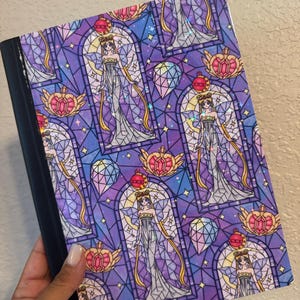 May include: A composition notebook featuring a repeating stained-glass style design. The design includes a figure in a long gown, surrounded by geometric shapes and celestial elements in shades of purple, blue, and pink. The notebook has a black spine.