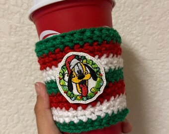 May include: A red disposable coffee cup with a white lid, held by a hand. The cup is wrapped in a handmade crochet cozy with red, white, and green stripes. A patch of Pluto in a Santa hat is on the cozy.