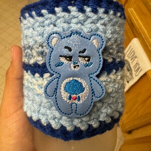 May include: Blue and white crocheted coffee cup cozy with a blue Care Bear character embroidered on the front. The Care Bear has a blue body, a white cloud on its belly, and a red heart on the cloud.