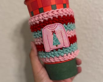 May include: A red and white plaid paper coffee cup with a white lid is wrapped in a handmade crochet cozy. The cozy is pink, red, and teal with a pink sweater applique featuring a green Christmas tree.