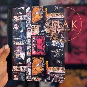 May include: Black notebook featuring a collage of anime-style illustrations in various colors. The cover includes characters, text, and graphic elements. The notebook is held by a person.